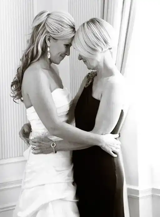 Bride and older woman embrace, sharing a tender, heartfelt moment in a warmly lit room.