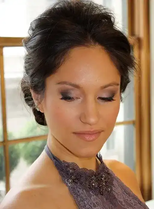Woman in a purple lace dress with an elegant updo, looking down and smiling subtly.