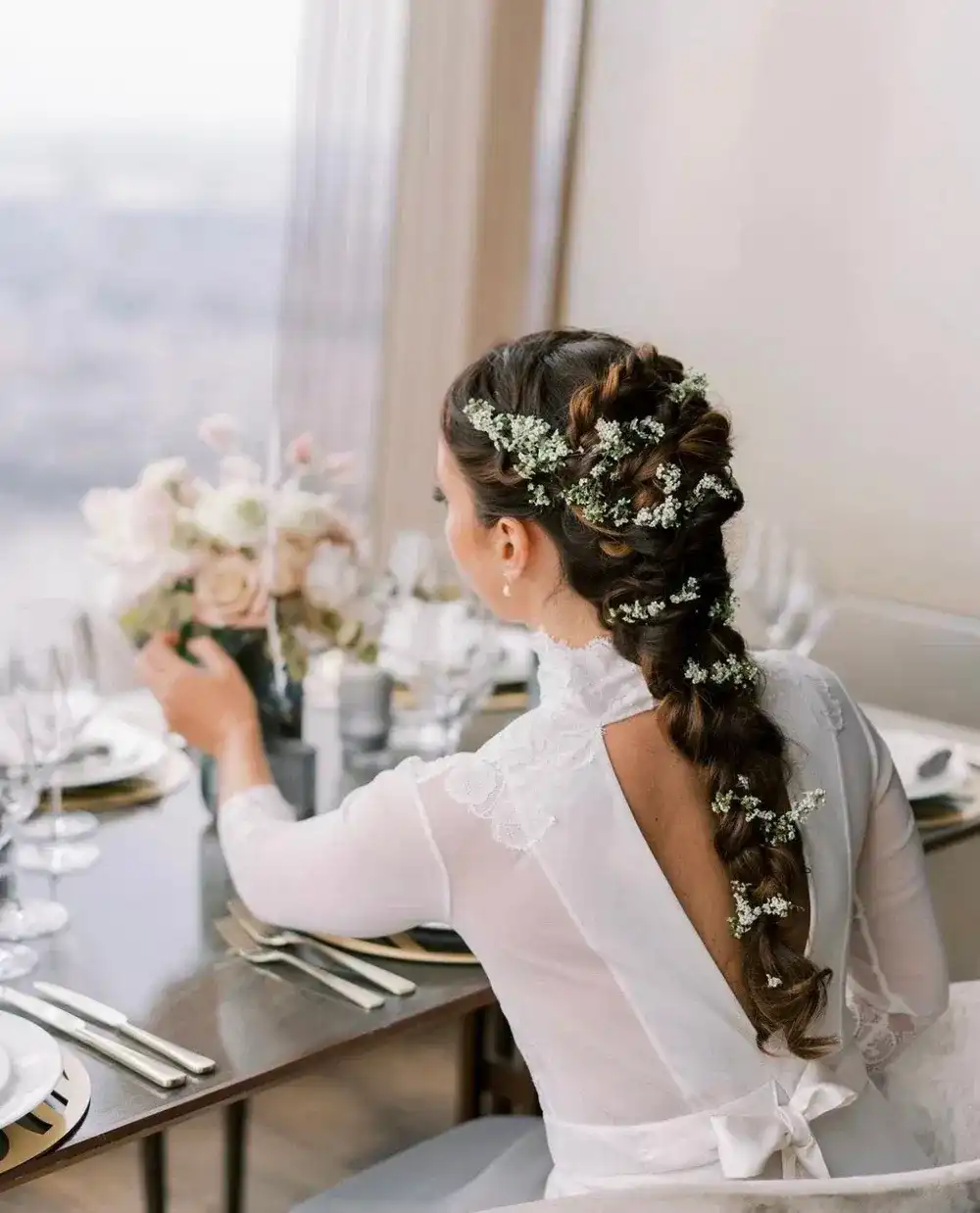 Bride with floral braid admires elegant table setting by a window.
