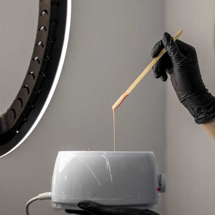 Warm wax being stirred with a spatula by a gloved hand in a salon setting.